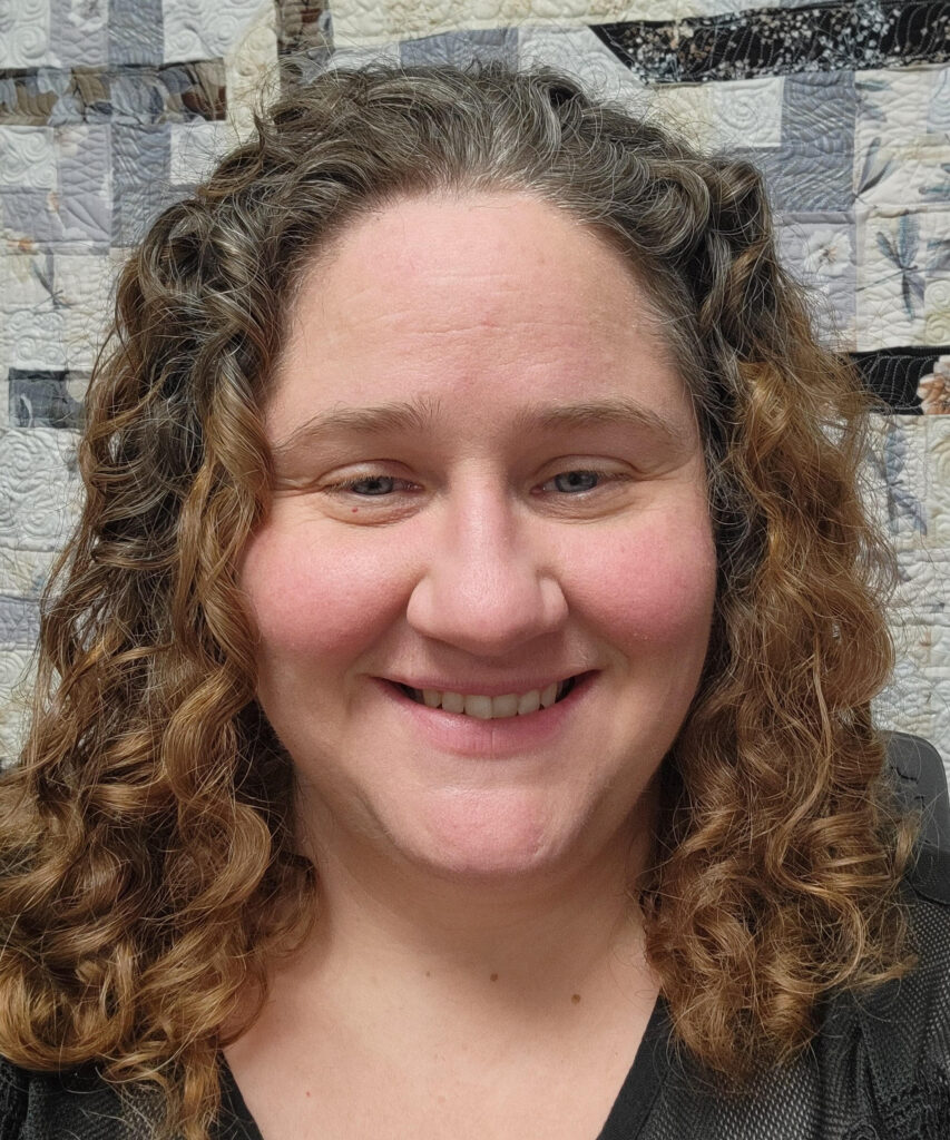 A portrait of a middle-aged woman smiling at the camera. She has long, curly brown hair with some gray streaks. The background behind her is a patchwork quilt with a pattern of geometric shapes and botanical elements in gray, white, and black tones.