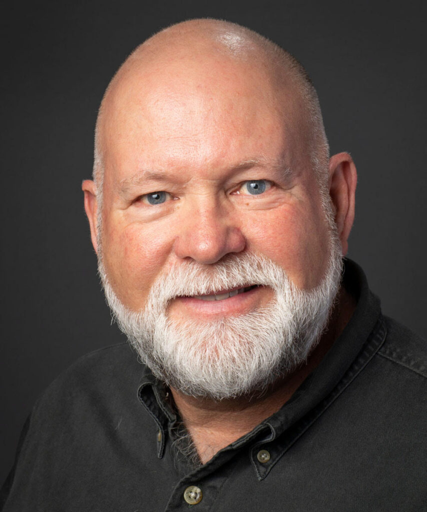 A studio portrait of a bald, middle-aged to older man, smiling at the camera. He has a full white beard and blue eyes. He is wearing a dark-colored (charcoal or dark gray) button-down shirt against a plain dark gray background.