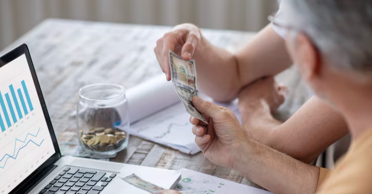 Close-up view of two people reviewing their finances at a table. A laptop displaying a financial chart is visible, and in the center, two hands are holding and counting one-hundred-dollar bills. On the table, there is a glass jar filled with coins and documents with pie charts.