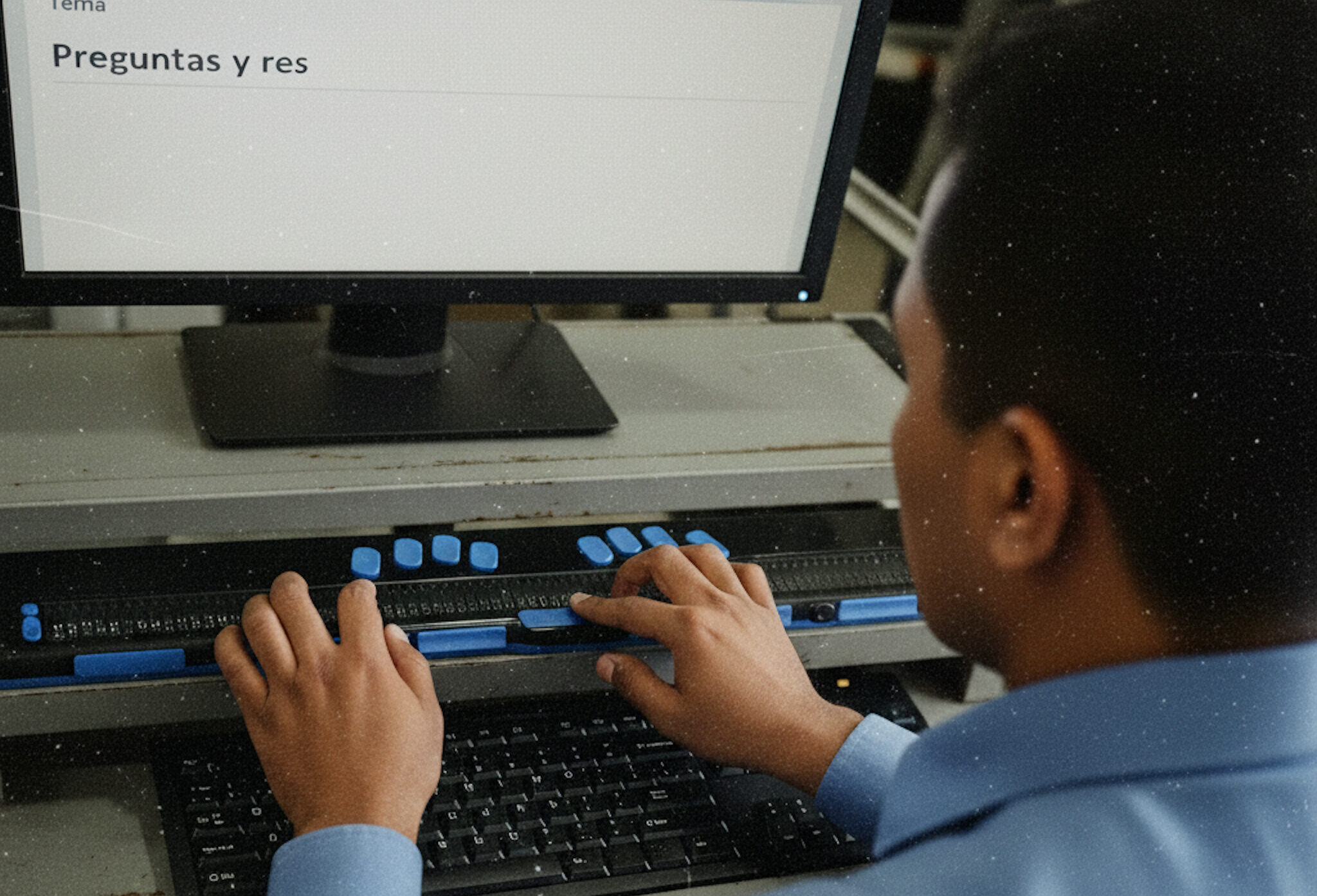 A man in a blue shirt is seated with his back to the camera in front of a computer, typing. He is using a standard QWERTY keyboard and a blue and black Braille display device placed just below the monitor. The computer screen shows a text document with the heading 'Preguntas y res...' (Questions and answers...).